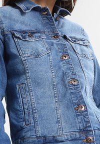 Chaqueta vaquera en azul claro con cierre de botones, dos bolsillos en el pecho y bolsillos laterales, con cuello clásico y detalles sutiles de costura.