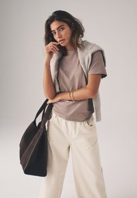 Brązowy t-shirt z krótkim rękawem w połączeniu z jasnobeżowymi spodniami o szerokich nogawkach. Szary sweter zarzucony na ramiona. Trzymana jest ciemnobrązowa torba typu tote.