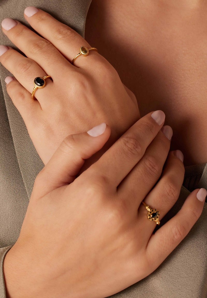 Zwei goldene Ringe an den Händen: Einer hat einen schwarzen ovalen Stein, der andere weist einen grünen ovalen Stein mit einem gemusterten Design auf. Neutrale Nagellackfarbe.