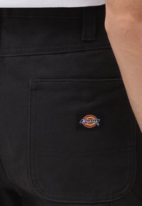 Primer plano del bolsillo de unos pantalones Dickies negros en la cadera de una persona con una etiqueta de marca visible.