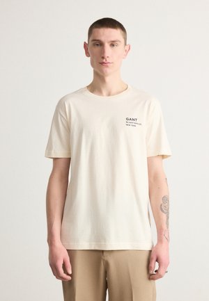 T-shirt en coton couleur crème à manches courtes et col rond, avec un logo imprimé noir sur la poitrine gauche.