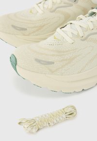 Beige Sportschuhe mit strukturiertem Obermaterial, grünen Sohlen und weißen Akzenten. Enthält eine gerollte gelb-weiße gestreifte Schuhschnürung darunter.
