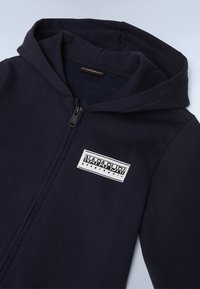 Felpa con zip color blu navy realizzata in morbido tessuto, con cappuccio, zip frontale e una patch rettangolare bianca con logo sul petto.
