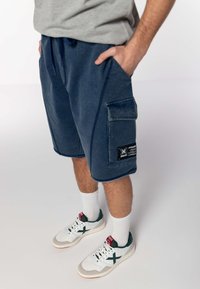 Dżinsowe szorty cargo w ciemnoniebieskim kolorze z tonującym przeszyciem, z boczną kieszenią i wszytą metką. W połączeniu z białymi i zielonymi trampkami.