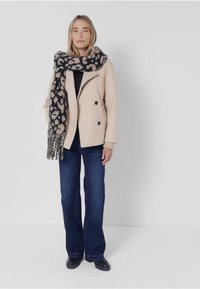 Beżowy płaszcz dwurzędowy, ciemny szalik w jasne cętki, czarna bluzka, ciemnoniebieskie dzwony i czarne buty. Gładki materiał, swobodny design.