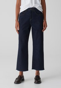 Jeans a gamba larga in denim blu scuro con orlo sfrangiato, cinque tasche e cuciture gialle a contrasto. Abbinati a mocassini neri.