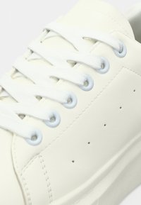 Tuvumā balts sporta apavu pāris ar auduma šņorēm, kas ievietotas metāla acīs uz gludas ādas virsmas.