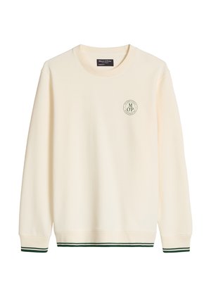 Jeune homme portant un sweat-shirt beige et une casquette, un short vert avec le numéro 67, des chaussettes blanches et des baskets blanches, debout sur un fond blanc uni.