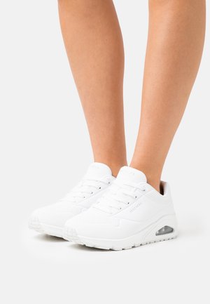 Beine, die weiße Skechers-Schnürschuhe mit sichtbar luftgepolsterten Sohlen auf weißem Hintergrund tragen.