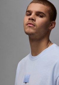 T-shirt in cotone azzurro chiaro con collo rotondo, presenta una toppa testurizzata sul petto. Maniche corte, vestibilità casual e una catena d'argento visibile.