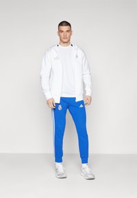 White zip-up hoodie with logo, paired with blue joggers featuring side stripes and a logo. White t-shirt underneath and gray sneakers.