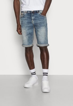 Hellblaue Jeansshorts mit umgeschlagenen Säumen, ausgestattet mit klassischen Taschen und Nahtdetails, kombiniert mit weißen Sneakers und gestreiften Socken.