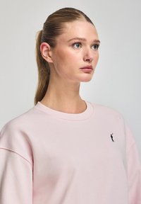 Light pink sweatshirt with drop shoulders, a ribbed neckline, and a small black logo on the left chest. Soft cotton fabric texture.