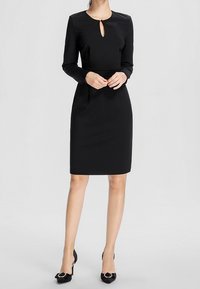 Femme portant une robe noire à manches longues, longueur genou, avec un décolleté en forme de trou de serrure, et des talons noirs, debout devant un fond clair uni.