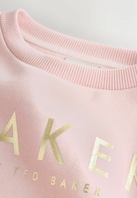 Różowa bluza z okrągłym dekoltem z złotym napisem "BAKER by Ted Baker" i ściągaczem, leżąca płasko na białej powierzchni.
