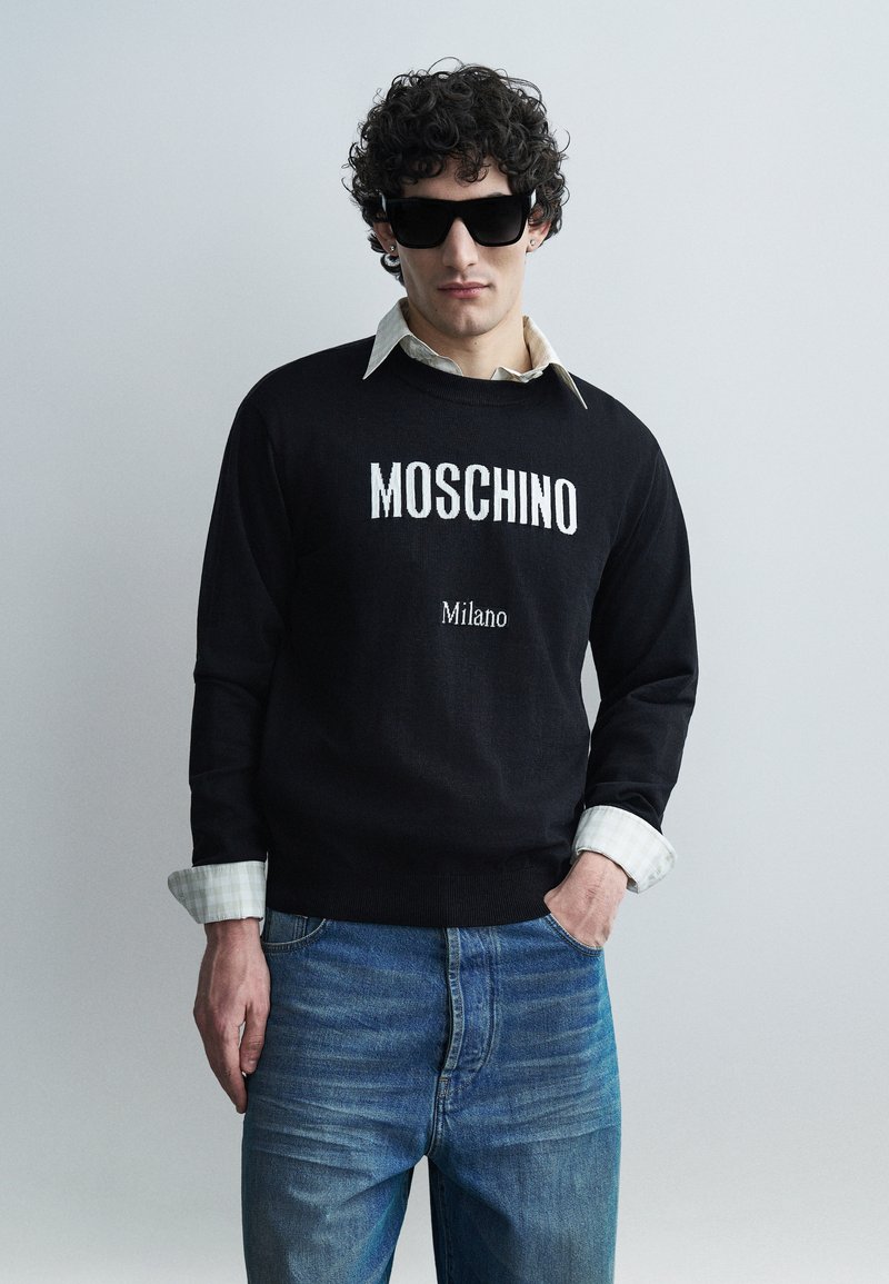 Homme aux cheveux bouclés portant un pull noir Moschino, une chemise blanche à col, un jean bleu et des lunettes de soleil noires sur un fond clair uni.