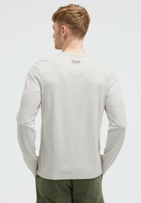 Helles graues Langarmshirt aus strukturiertem Stoff, runder Ausschnitt und ein kleines Etikett auf der Rückseite. Kombiniert mit olivgrünen Hosen.