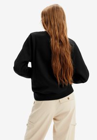 Sudadera negra con cuello redondo, hombros caídos y puños acanalados, combinada con pantalones cargo beige que tienen bolsillos laterales y detalles de botones.