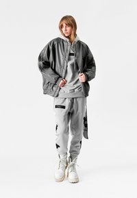Chaqueta bomber de cuero gris sobre una sudadera con capucha gris claro y pantalones de chándal a juego con acentos y parches negros, combinados con botas blancas.