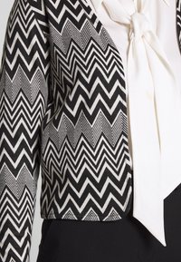 Black and white cropped jacket with a zigzag pattern, featuring a light-colored tie detail and a smooth texture.