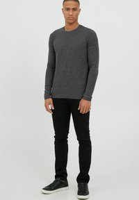 Graues Langarm-T-Shirt mit Rundhalsausschnitt, körperbetonter Form und kontrastierendem braunem Besatz, kombiniert mit schwarzen Slim-Fit-Jeans und schwarzen Turnschuhen.