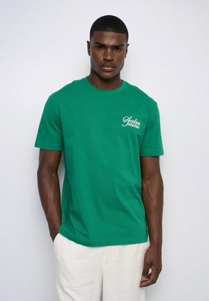 Homme portant un t-shirt vert manches courtes avec le texte blanc « Salsa Jeans » et un pantalon blanc, debout devant un fond clair uni.