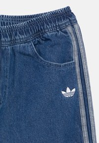 Μπλε τζιν φόρμα με ελαστική ζώνη στη μέση, πλευρικές ρίγες και άσπρο κεντητό λογότυπο στην αριστερή τσέπη. Μαλακή υφή, casual σχεδιασμός.