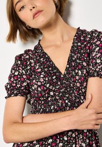 Musta kukallinen mekko lyhyillä puffi-hihoilla. Ominaisuuksiin kuuluu hyrretetty yläosa ja syvä V-pääntie. Kuvioita ovat vaaleanpunaiset, valkoiset ja keltaiset kukat.