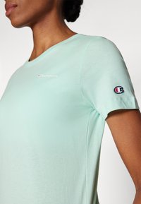 Miętowo-zielona koszulka z krótkim rękawem wykonana z miękkiego materiału, z okrągłym dekoltem i małym wyszywanym logo Champion na lewej piersi oraz rękawie.