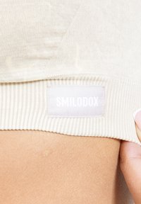 Vaaleanbeige lyhyt toppi, jossa on ribbikuviointi, ja valkoinen SMILODOX-tunnus ommeltuna helmaan. Sujuva pinta, istuva malli.