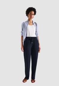 Marineblauwe relaxte broek met een trekkoord in de taille, gecombineerd met een witte tanktop en een blauw-wit gestreepte blouse. Bruine sandalen.