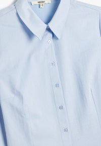 Camisa de botones azul claro con cuello puntiagudo, que presenta una textura de espiga y seis botones a juego en la parte delantera.