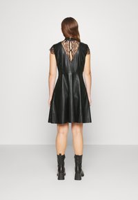 Femme portant une robe en cuir noire avec un détail en dentelle dans le dos et des bottines noires, debout de dos devant un fond clair uni.