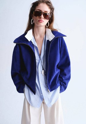 Kobieta nosząca oversize niebieską kurtkę na paskowanej koszuli, białe spodnie, duże kolczyki koła, okulary przeciwsłoneczne i długi naszyjnik z zawieszką.