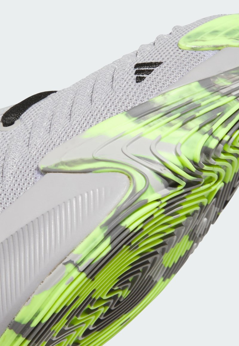 Chaussure de sport en mesh gris clair avec une surface texturée. Dispose d'une semelle à motifs verts et noirs et d'un logo noir accentué sur le côté.