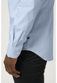 Ljust blå skjorta med en figurnära design, långa ärmar, knappmanchetter och en sidomärkning som anger storlek. Svarta byxor syns i botten.