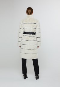 Bílé umělé kožich s vodorovnými černými pruhy a černým páskem v pase. Obsahuje vysoký límec a dlouhé rukávy.