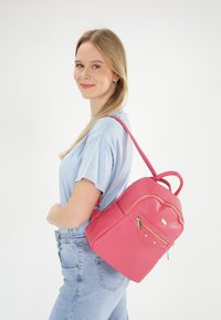 Sac en cuir texturé rose avec une poche frontale à fermeture éclair, quincaillerie dorée et poignées supérieures doubles ; conçu pour être porté sur une épaule.