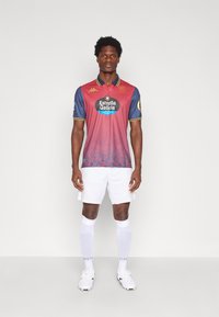 Sarkans un zils futbola krekls ar apkakli, kuram ir "Estrella Galicia" logo, savienots ar baltām šortiem un zeķēm. Redzami melni apavi.