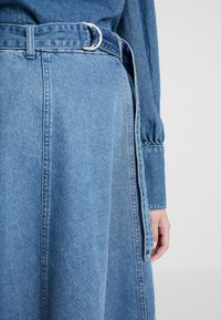 Denimklänning med blå färg, bälte i midjan med en silverring och lös passform. Texturen är mjuk med synliga sömnadsdetaljer.