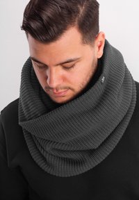 Dark gray knitted infinity scarf with a textured pattern, featuring a subtle logo accent on the edge. Worn loosely around the neck.
