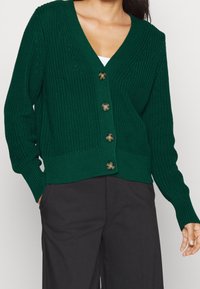 Grön stickad cardigan med V-ringning, fem stora runda knappar, ribbade ärmslut och nederkant; har ett texturerat mönster. Bärs över mörka byxor.