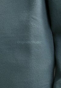 Texturierter grüner Stoffauschnitt mit einem weichen Strickmuster, das den eingeprägten Text "Originals Studio" in einem dezenten Design zeigt.