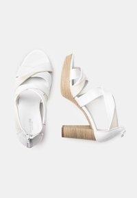 Sandales à talons hauts blanches avec des bretelles croisées, un bout ouvert et un talon bloc texturé de couleur marron clair. Fini lisse avec fermeture éclair.