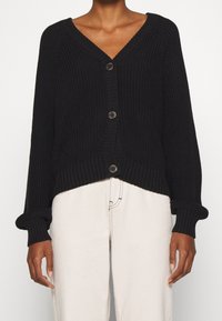 Persona vestida con un cárdigan de punto negro con botones y puños enrollados, y pantalones de color blanco roto, mostrada de cuello a muslos.