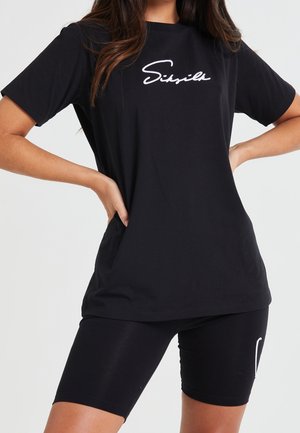 Kobieta w czarnej koszulce z białym logo "SikSilk" i dopasowanych czarnych szortach rowerowych, dłonie spoczywają na biodrach.