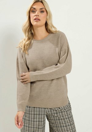 Blonde Frau trägt einen beigen Strickpullover und schwarz-weiß karierte Hose, steht vor einem einfarbigen hellen Hintergrund.