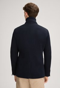 Blazer in misto lana blu navy con alto colletto, spalle strutturate e vestibilità su misura. Presenta una cucitura centrale e tasche laterali. Texture liscia.