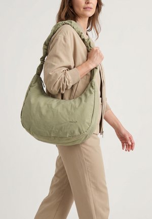 Vrouw in beige outfit draagt een grote lichtgroene schoudertas met een gewatteerde band, lopend tegen een eenvoudige achtergrond.