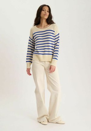 Femme debout, portant un pull beige avec des rayures horizontales bleues, un pantalon large crème et des chaussures crème sur un fond uni.